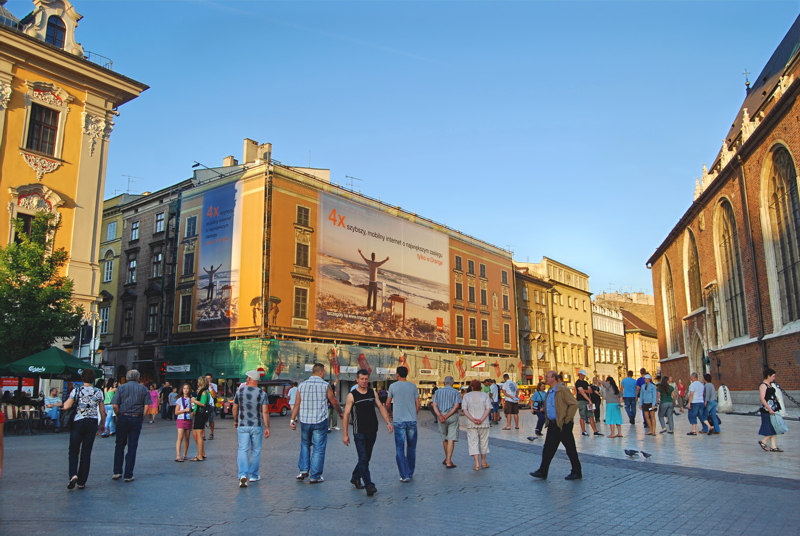 15 - The bustling atmosphere of the Main Market Square in Kraków, featuring pedestrians strolling past historic architecture and a large building facade under a clear blue sky